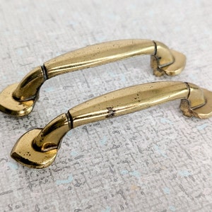 May include: Two gold-toned metal drawer pulls with a curved design. The pulls have a smooth, polished finish.