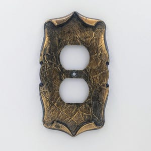 May include: Ornate, vintage-style electrical outlet cover with a textured, bronze-coloured finish. The cover has a decorative, shield-like shape with two circular openings for the outlets and a central screw hole.