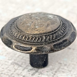 May include: A round, antique-style cabinet knob with a raised, textured design. The knob is made of metal and has a dark bronze finish.