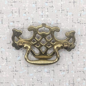 May include: An ornate brass drawer pull with a decorative design. The pull has a central diamond pattern and a curved handle. The hardware is a warm gold colour, suitable for antique furniture restoration or DIY projects.