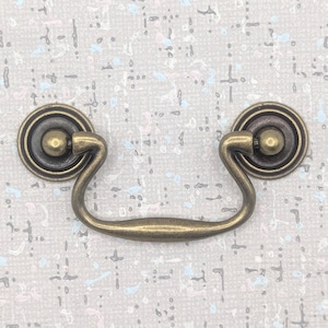 以下が含まれることがあります： カーブしたハンドルと円形のバックプレートを備えたアンティーク真鍮仕上げの引き出しプル。 このハードウェアは、キャビネットや家具に適したクラシックなデザインが特徴です。 プルは金属製で、ヴィンテージ感があります。