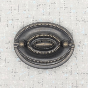 May include: An antique-style oval drawer pull with a dark bronze finish. The pull features a detailed design with a central oval and beaded edges, and small ball-shaped accents on each end. A classic hardware piece.