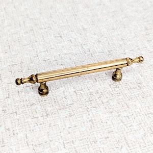 May include: A gold-toned metal cabinet pull with a long, straight bar and rounded ends. The pull has a slightly aged, antique look.