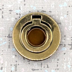 May include: A gold-colored phone ring holder with a circular design. The ring holder has a round base with a raised, circular ring in the center. The ring is designed to rotate and fold flat against the base. The background is a speckled gray.