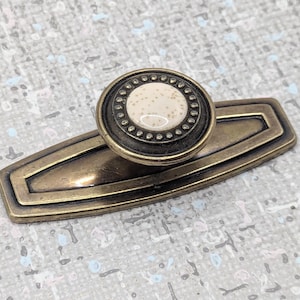 Peut inclure: Tirette de meuble en laiton antique avec un bouton rond orné d'un centre crème moucheté et d'une bordure décorative. La tirette a une plaque arrière rectangulaire au design élégant. Une pièce de quincaillerie classique.