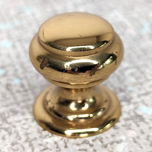 May include: A gold-coloured cabinet knob with a rounded top and a tiered base. The knob shows signs of wear, with small areas of discolouration. The background is a light, textured surface.