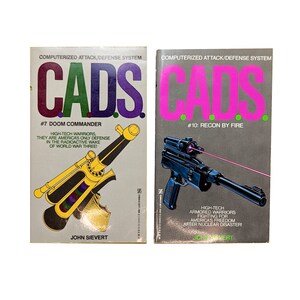May include: Two vintage paperback books from the CADS series. The first book, "#7 Doom Commander," features a yellow and black weapon illustration. The second book, "#10: Recon by Fire," has a pink laser gun illustration. Both books have the text "COMPUTERIZED ATTACK/DEFENSE SYSTEM."