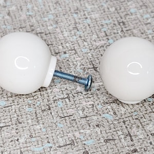 May include: Two white, spherical cabinet knobs with a glossy finish. Each knob has a metal screw for installation. The knobs are shown on a textured, light grey surface.