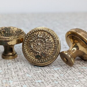 May include: Three antique brass cabinet knobs with ornate floral designs. The knobs have a warm, aged patina, showcasing intricate detailing and a vintage aesthetic. Each knob features a round face and a short, cylindrical base.