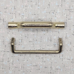 May include: Two gold-coloured metal hardware pieces. One is a straight bar with rounded ends, and the other is a U-shaped piece. Both pieces are on a light-coloured, speckled background.
