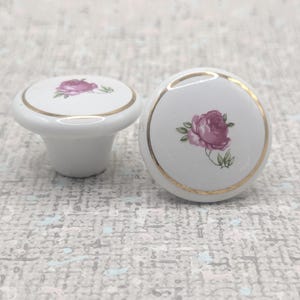 May include: Two white ceramic cabinet knobs with a gold trim and a pink rose design. One knob is round, and the other is a mushroom shape. The knobs are likely for a drawer or cabinet.