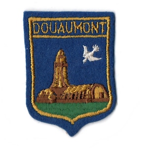 May include: Embroidered patch with a blue background and gold trim. The patch features the word "DOUAUMONT" in gold, a brown and gold building, a white bird, and green grass.