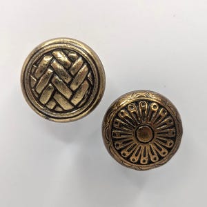 May include: Two gold-toned metal cabinet knobs with decorative designs. One knob has a woven pattern, and the other has a sunburst design.