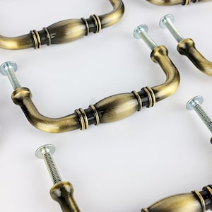 May include: Antique brass cabinet hardware pulls with a decorative design. The pulls have a curved shape and are attached to screws.