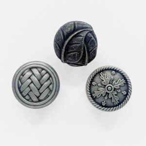 May include: Three silver-toned cabinet knobs with different designs. One knob features a woven pattern, another has a leaf design, and the third has a stylized oak leaf design.