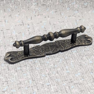 May include: Antique bronze-toned cabinet pull with a decorative design. The pull features a textured backplate and a detailed handle with a central rope-like element. The handle is supported by two posts.