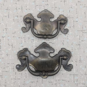 以下が含まれることがあります： 2つのアンティーク真鍮引き出しプル。各プルは、湾曲したハンドルと装飾的な装飾が施された装飾的なバックプレートを備えています。プルは金属製で、変色した古い外観をしています。明るい灰色の背景に配置されています。