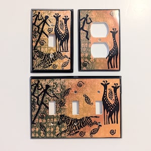 Peut inclure: Ensemble de trois plaques de prise décoratives sur le thème de la savane africaine. Les plaques sont beiges avec des contours noirs et représentent des girafes, des zèbres et des figures humaines stylisées.