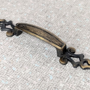 Peut inclure: Une poignée de tiroir décorative en or antique avec des volutes ornées aux extrémités. La poignée a une finition texturée et vieillie.