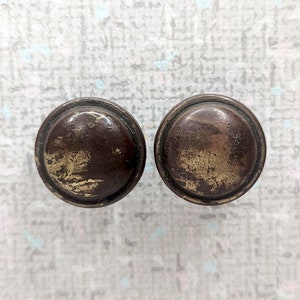 May include: A pair of antique, round cabinet knobs with a dark brown finish and a distressed appearance. The knobs have a slightly domed shape and a brassy, golden hue in areas, showing signs of age and wear.