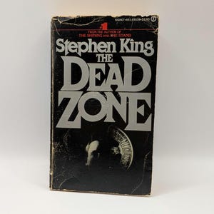 May include: A mass market paperback book titled The Dead Zone by Stephen King. The cover is black with large silver lettering for the title and author name. A small graphic of a roulette wheel is visible near the bottom center of the cover.