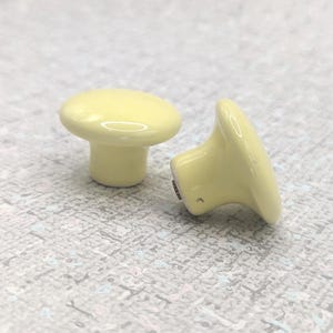 Peut inclure: Deux boutons de meuble en céramique jaune pâle en forme de champignon. Les boutons ont un dessus arrondi et une base cylindrique courte. Les boutons sont probablement pour un tiroir ou une armoire. Le fond est une surface mouchetée grise et blanche.
