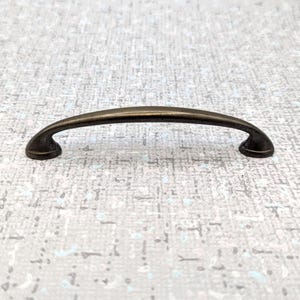 May include: A bronze-colored cabinet pull with a curved handle and rounded ends. The hardware is designed for easy gripping and is likely used for drawers or cabinet doors. The pull is made of metal and has a vintage or antique appearance.