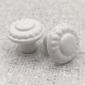 以下が含まれることがあります： 白のセラミック製キャビネットノブが2つ。各ノブは、装飾的なエッジが付いた丸くわずかにドーム型のトップを備えています。ノブは、明るい色の斑点のある背景に対して配置されています。ノブはおそらく家具用です。