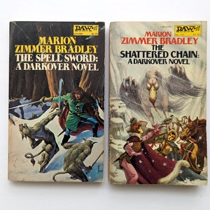 May include: Two paperback fantasy novels with covers featuring illustrations of a man fighting a monster and a group of people running from a large bird-like creature. The books are both titled "Darkover Novels" and feature the author Marion Zimmer Bradley.