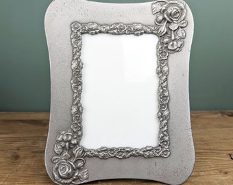May include: A silver-toned picture frame with a decorative floral design. The frame has a slightly curved shape and a raised border.