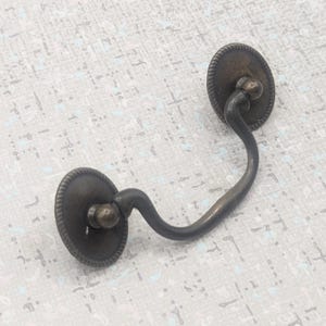 以下が含まれることがあります： アンティークブロンズの引き出し取っ手。曲線的なハンドルと、装飾的な丸いバックプレートが2つ付いています。バックプレートにはロープのような縁取りがあります。キャビネットや引き出し用に設計された金具です。