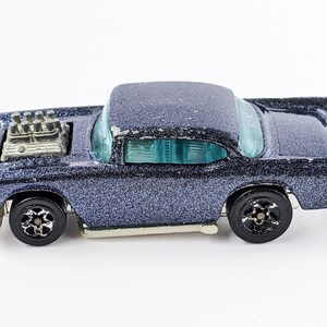 May include: A vintage die-cast model car in a dark blue glitter finish. The classic car features a prominent engine detail and black wheels. The windows are a light blue color. The car is a collectible toy.
