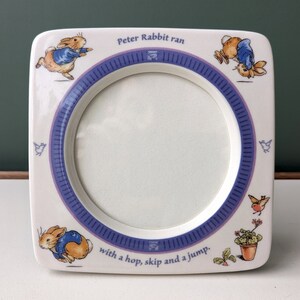 May include: A white ceramic picture frame with a blue and purple border. The frame features illustrations of Peter Rabbit running with the text "Peter Rabbit ran with a hop, skip and a jump."