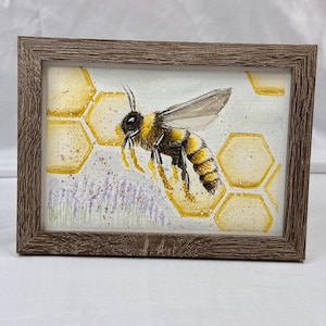 May include: Watercolor painting of a bee with yellow and black stripes, wings extended, hovering near a honeycomb. The artwork is framed in a brown wooden frame, with a white background and lavender flowers.