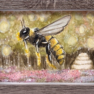 May include: Watercolor painting of a bee in flight, framed in a rustic wooden frame. The bee is detailed with black and yellow stripes, wings spread, and pollen visible. The background features a soft, blurred landscape with a beehive.