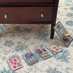 May include: A dark brown wooden miniature chest of drawers with two small knobs. In front are five rectangular cards featuring vintage-style illustrations and attached embellishments, including pink beads, a gold spool, and white buttons.