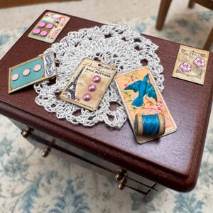 May include: A collection of miniature sewing supplies including four small rectangular cards with vintage-style graphics, buttons, and a tiny wooden spool with blue thread, all arranged on a white crocheted doily. The cards feature illustrations of the Eiffel Tower, a blue bird, and floral patterns.