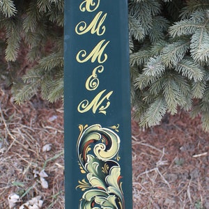 May include: A dark green wooden sign with gold lettering and floral scrollwork. The sign has the letters "A E K M M E N" in a decorative font. The bottom of the sign features a detailed floral design in shades of green, yellow, and red.