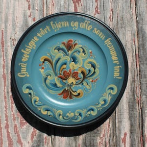 May include: A round decorative plate with a black rim and a light blue center. The center features hand-painted floral scrollwork in red, yellow, and blue. The text "Gud velsigne vårt hjem og alle som kommer inn!" is written in gold lettering along the top.