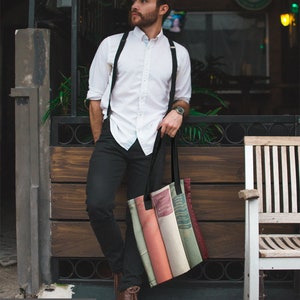 May include: A black tote bag featuring a print of vintage books in various colors. The bag has black handles and is held by a person wearing a white shirt, black pants, and brown boots. The image includes the text "Tiffany Dawn Smith PHOTOGRAPHY".