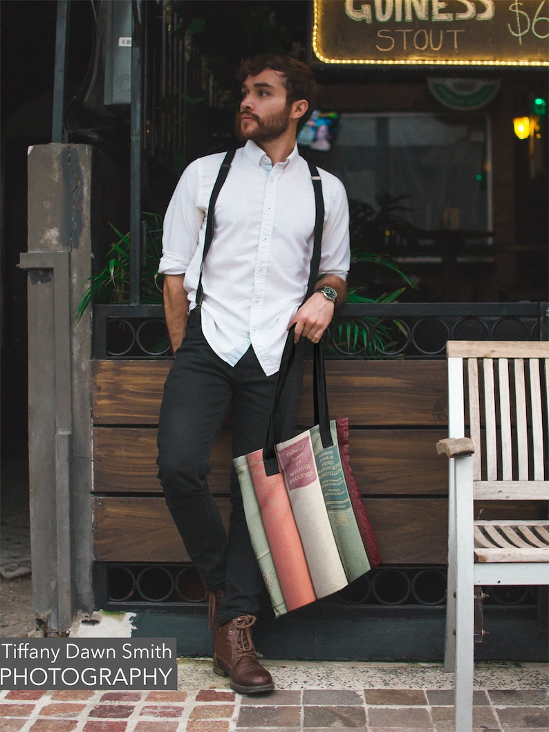 May include: A black tote bag featuring a print of vintage books in various colors. The bag has black handles and is held by a person wearing a white shirt, black pants, and brown boots. The image includes the text "Tiffany Dawn Smith PHOTOGRAPHY".