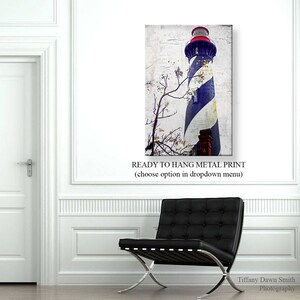 May include: Metal print of a lighthouse with blue and white stripes, a red top, and a brick base. The print has a distressed, vintage look. The text "READY TO HANG METAL PRINT" is at the bottom. A black leather chair is in the foreground.