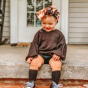 May include: A young child wearing a black sweater with puffed sleeves, black knee-high socks, and black and white sneakers with blue laces. The child is wearing a floral headband with a large bow. The child is sitting on a set of steps.