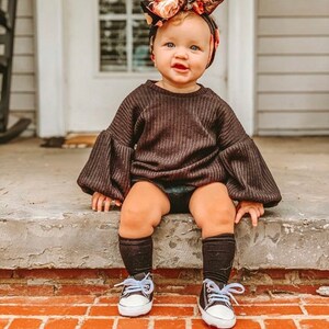 May include: A baby girl wearing a black sweater with puffed sleeves, black socks, and white sneakers. She is sitting on a brick step and wearing a floral headband.