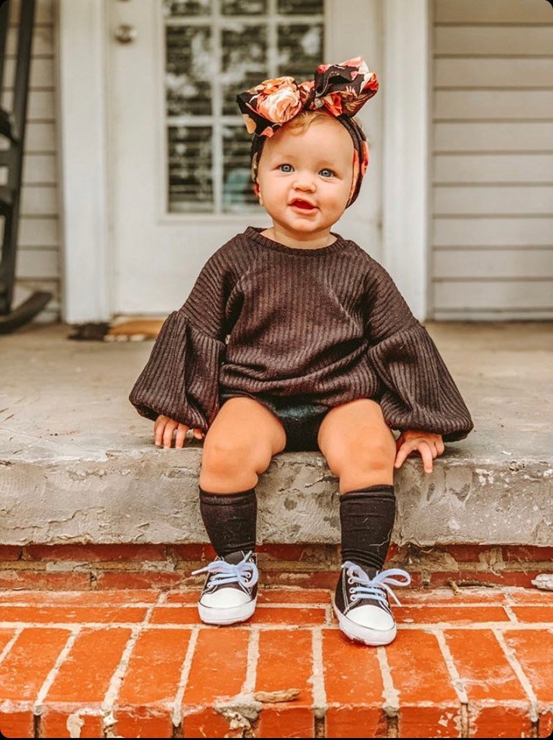 May include: A baby girl wearing a black sweater with puffed sleeves, black socks, and white sneakers. She is sitting on a brick step and wearing a floral headband.