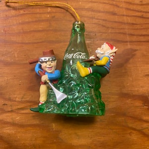 May include: A green glass Coca-Cola bottle Christmas ornament with two elves. One elf is holding a pickaxe and the other is holding a bottle of Coca-Cola.