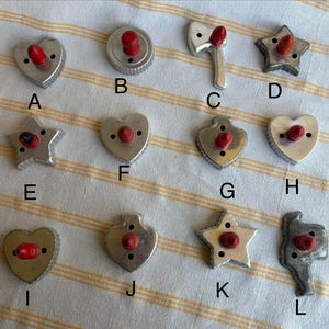 May include: A collection of vintage metal cookie cutters in various shapes, including hearts, stars, a circle, an axe, and a Texas-shaped cutter. Each cutter has a red knob. The cutters are arranged on a striped, cream-colored surface.