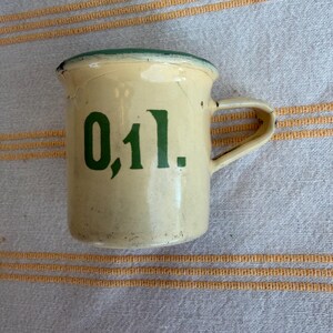 May include: A small, vintage cream-colored enamel mug with a green rim and handle. The mug features the number "0,1 l." in green lettering on the front. The mug has a slightly distressed appearance.