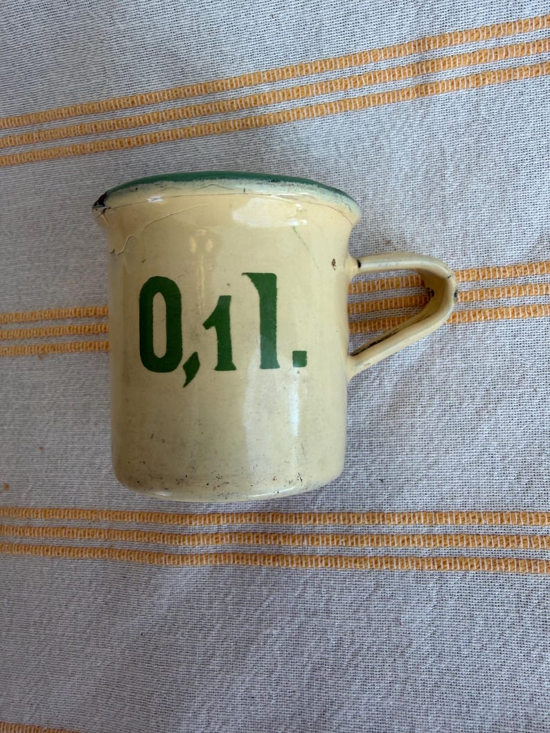 May include: A small, vintage cream-colored enamel mug with a green rim and handle. The mug features the number "0,1 l." in green lettering on the front. The mug has a slightly distressed appearance.