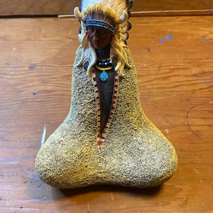 Puede incluir: Una figurita de un jefe nativo americano con un tocado de plumas y un collar de cuentas. El jefe está de pie sobre una base texturizada que se asemeja a un montículo de tierra.